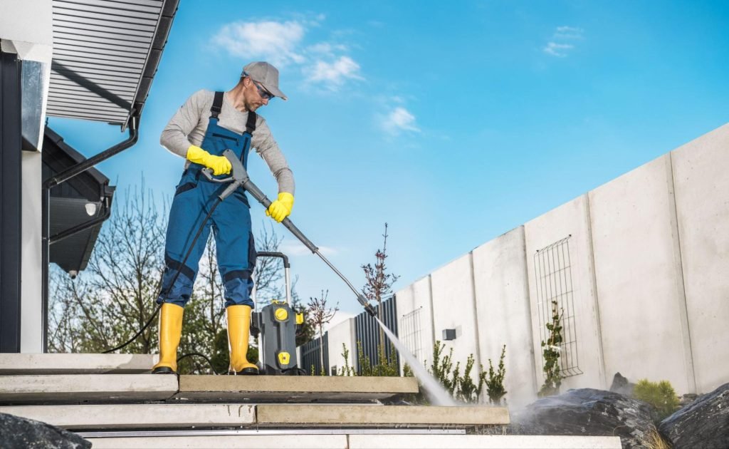A professional in rubber boots and personal protective equipment cleans the steps and rock features outside a business using a pressure washer
