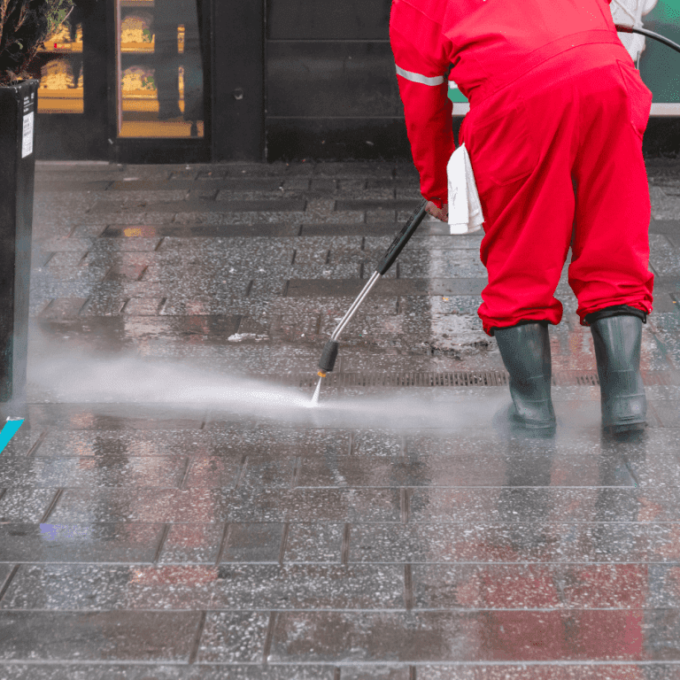 A professional in rubber boots and a red jumpsuit pressure washes a paved outdoor walkway
